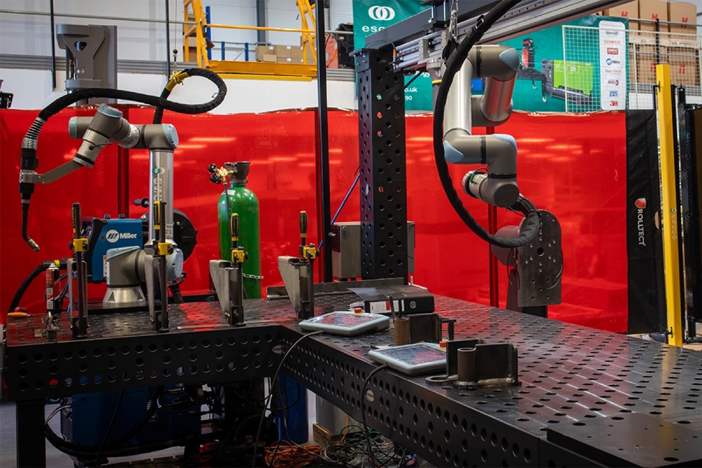 A collaborative robot arm MIG welding a steel part clamped to a Siegmund fixturing table, arc lit